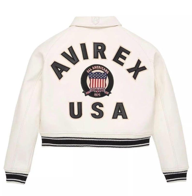Puede incluir: Chaqueta color crema con ribetes a rayas blancas y negras. La parte trasera presenta la palabra "AVIREX" sobre un parche circular con un dise&ntilde;o de la bandera estadounidense y la palabra "USA" debajo. La chaqueta tiene un estilo bomber cl&aacute;sico.