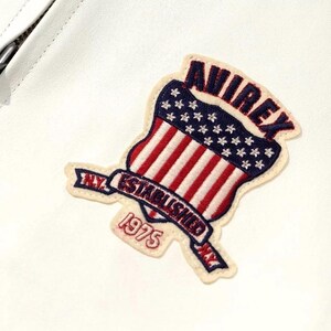 Puede incluir: Parche bordado con un dise&ntilde;o de escudo con rayas rojas y blancas, estrellas azules y la palabra "AVIREX". Una pancarta debajo dice "N.Y. ESTABLISHED 1975". El parche est&aacute; sobre un fondo blanco.