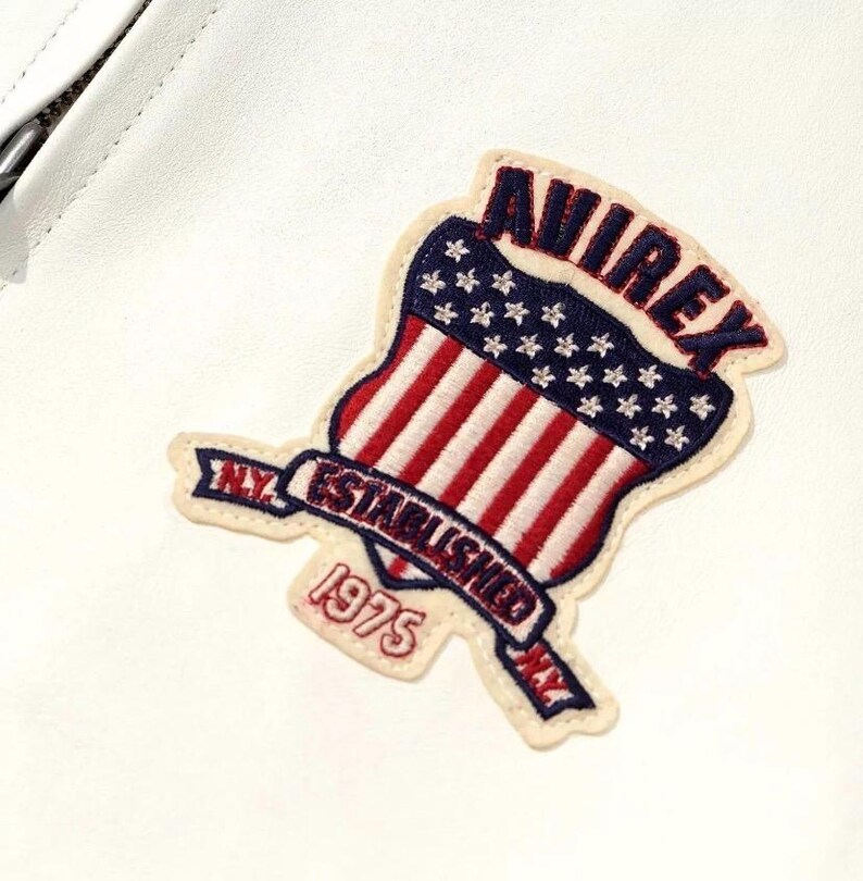 Puede incluir: Parche bordado con un dise&ntilde;o de escudo con rayas rojas y blancas, estrellas azules y la palabra "AVIREX". Una pancarta debajo dice "N.Y. ESTABLISHED 1975". El parche est&aacute; sobre un fondo blanco.