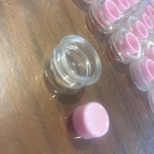 May include: A selection of small, clear plastic jars with pink lids. The jars are arranged on a wooden surface. The lids are a solid, light pink colour. The jars appear to be empty and ready for filling.