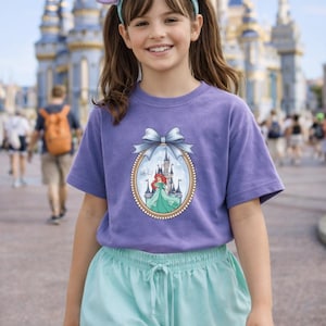 Peut inclure: Une personne porte un t-shirt violet avec une image encadr&eacute;e d'une princesse et d'un ch&acirc;teau. La personne porte &eacute;galement un bandeau avec des oreilles de souris et un n&oelig;ud, ainsi qu'un short bleu clair. En arri&egrave;re-plan, un ch&acirc;teau.