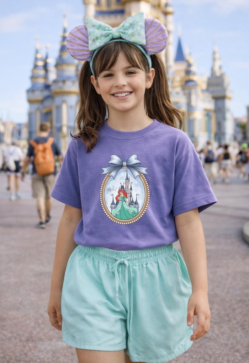 Peut inclure: Une personne porte un t-shirt violet avec une image encadr&eacute;e d'une princesse et d'un ch&acirc;teau. La personne porte &eacute;galement un bandeau avec des oreilles de souris et un n&oelig;ud, ainsi qu'un short bleu clair. En arri&egrave;re-plan, un ch&acirc;teau.