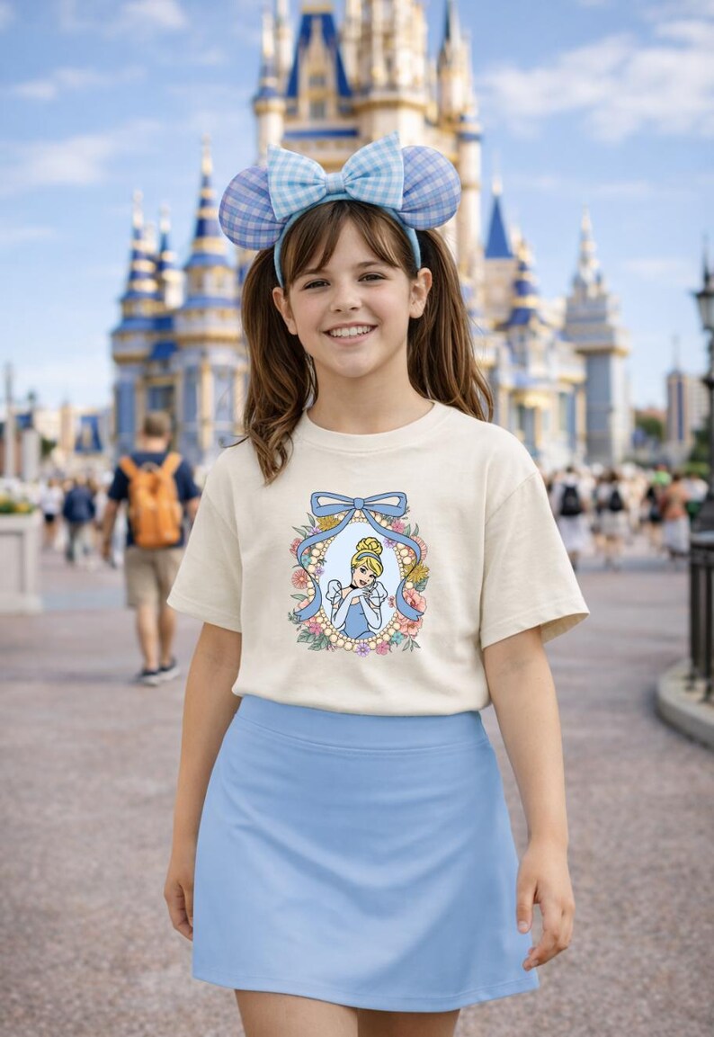 May include: A young person wearing a cream-colored t-shirt with a Cinderella graphic, a light blue skirt, and a blue and white plaid bow headband. The background features a castle and a crowd of people.