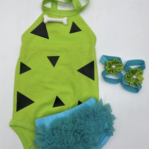 May include: A lime green baby outfit with black triangle accents and a bone-shaped embellishment. Includes a pair of blue ruffled bloomers and two blue sandals with green flower accents. The outfit is designed for a baby.