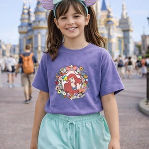 Peut inclure: Une jeune personne porte un t-shirt violet avec une image encadr&eacute;e de fleurs d'Ariel de La Petite Sir&egrave;ne. La personne porte &eacute;galement un bandeau avec des oreilles de souris et un n&oelig;ud, et un short bleu clair. Un ch&acirc;teau est en arri&egrave;re-plan.