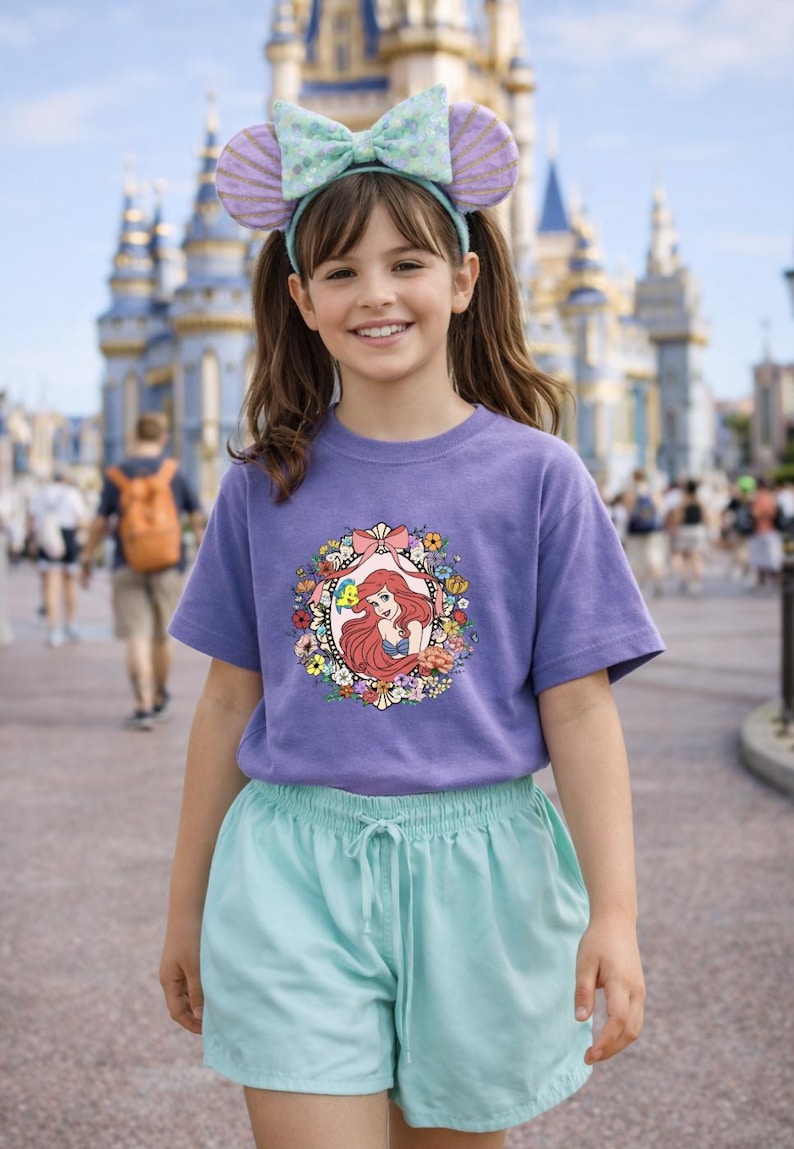 Peut inclure: Une jeune personne porte un t-shirt violet avec une image encadr&eacute;e de fleurs d'Ariel de La Petite Sir&egrave;ne. La personne porte &eacute;galement un bandeau avec des oreilles de souris et un n&oelig;ud, et un short bleu clair. Un ch&acirc;teau est en arri&egrave;re-plan.