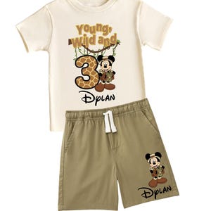 May include: A two-piece children's outfit featuring a cream-colored t-shirt and khaki shorts. The t-shirt has the text "Young, Wild and 3" with a giraffe-patterned number 3 and a Mickey Mouse graphic. The shorts have a Mickey Mouse graphic and the name "Dylan".