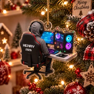 May include: A clear acrylic Christmas tree ornament featuring a person in a grey hoodie and headphones sitting in a black gaming chair. The chair back displays the text HENRY 2026. The person faces a computer monitor, keyboard, and tower with glowing lights.