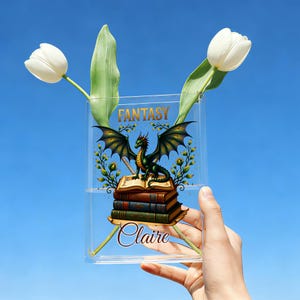 Op de afbeelding: Een heldere acrylvaas met een fantasiethema, met een groene draak op een stapel boeken. Het woord "FANTASY" staat bovenaan en "Claire" staat onderaan. Twee witte tulpen zijn in de vaas geplaatst.