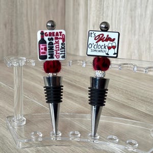 May include: Two wine bottle stoppers with decorative tops. One has the text "Great minds drink alike" and a wine bottle graphic. The other says "It's Wine O'clock Somewhere" with a wine glass. Both have a red and black pom-pom.