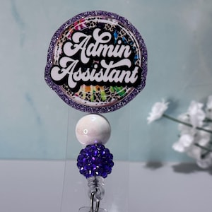 May include: A round badge reel with the words "Admin Assistant" in white script. The badge has a purple glitter border and a colorful tie-dye design. It features a white bead, a cluster of purple beads, and a clear plastic clip.