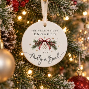 May include: A round white ceramic ornament hanging by a cream ribbon. It features the text THE YEAR WE GOT ENGAGED, a red bow with pine branches and berries, a small heart charm, the year 2026, and the names Molly & Brad in black script.