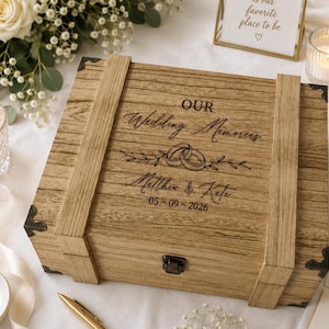 May include: A rectangular wooden keepsake box with a hinged lid and dark metal corner accents. The lid features the engraved text "OUR Wedding Memories" above an illustration of two interlocking rings, the names "Matthew & Kate," and the date "05 ~ 09 ~ 2026."
