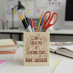 May include: A light-coloured wooden pencil holder featuring brown engraved illustrations of school supplies and the text "MY LITTLE POT OF TEACHING STUFF MISS. JACKSON". The holder contains various coloured pencils, pens, and red scissors.