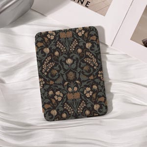 May include: A rectangular e-reader case featuring a dark black background with a repeating botanical pattern of muted green leaves, tan flower buds, and small beige blossoms arranged in a symmetrical, intricate, and dense floral design.