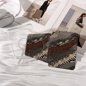 May include: A clear phone case and a patterned phone case with a mix of animal prints, including zebra and leopard patterns, in shades of brown, gray, and beige. The phone case is lying on a white surface next to a pair of pink headphones and a magazine.