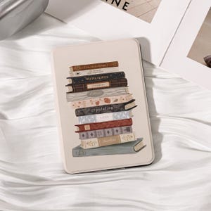 May include: A cream e-reader case with a design of stacked books, including titles such as "Midnights" and "Folklore." The books are in shades of brown, blue, and red. The case is rectangular and has a smooth finish.