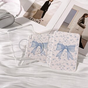 May include: A clear phone case and a floral patterned phone wallet with a light blue bow design. The wallet has a matching pop-up grip. The background includes fashion magazines and a pair of headphones.
