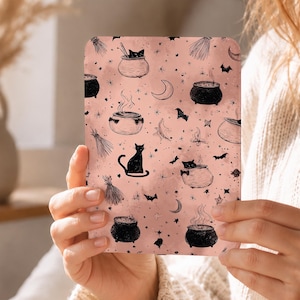 May include: A pink rectangular card featuring a repeating pattern of black witch cauldrons, cats, bats, brooms, moons, and stars. The card is held by a person.