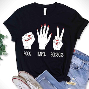 May include: Black t-shirt with a graphic of three hand gestures: rock, paper, and scissors. The hands are white with red nail polish. The words "ROCK", "PAPER", and "SCISSORS" are printed below the hands in white.