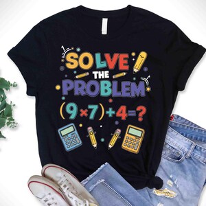 May include: Black t-shirt featuring the phrase "SOLVE THE PROBLEM" in vibrant colours, a maths equation, and pencil and calculator illustrations. The shirt is styled with blue jeans and white trainers.