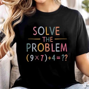 May include: Black t-shirt with the text "SOLVE THE PROBLEM (9x7)+4=??" in colorful letters. The shirt is a crew neck and has short sleeves. The text is in a playful font.