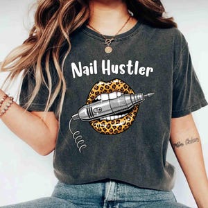 May include: Dark gray t-shirt with the text "Nail Hustler" above a graphic of lips with a leopard print design holding a silver nail drill. The shirt is a casual style.