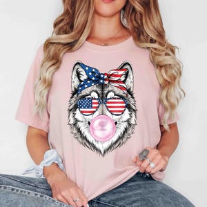 May include: A light pink t-shirt with a wolf graphic. The wolf is wearing sunglasses with an American flag design and a matching headband. A pink bubble gum bubble is visible. The graphic is centered on the shirt.