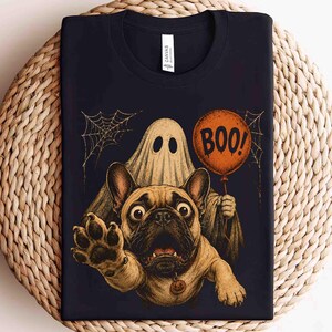 Może przedstawiać: Czarny t-shirt z grafiką przedstawiającą beżowego buldoga francuskiego z zaskoczoną miną, wyciągającego łapę do przodu. Za psem znajduje się duch trzymający pomarańczowy balon z napisem BOO! oraz wzory pajęczyn.