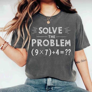 May include: Charcoal grey t-shirt featuring the phrase "SOLVE THE PROBLEM" and the mathematical equation (9 x 7) + 4 = ??. The white text is bold and clear. A relaxed fit, suitable for casual wear.