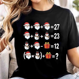 May include: Black t-shirt featuring a Christmas-themed mathematical puzzle. The design includes illustrations of Santa Claus, snowmen, and gift boxes, with an incomplete equation asking for the solution. The text is in white.