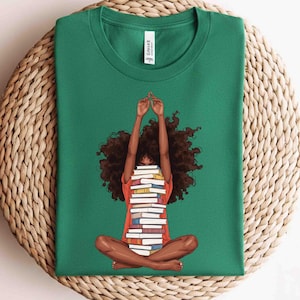 May include: A forest green t-shirt featuring a graphic illustration of a person with dark curly hair sitting in a cross-legged position, holding a tall stack of books in front of their face with both arms raised high above their head.