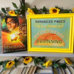 May include: A bookshelf features a book titled "Dawn of Vengeance" and a framed cross-stitch with the words "Darkness Fades, The Sun Comes Out, and there's a new Beginning." Yellow sunflowers and greenery decorate the shelf.