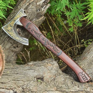 May include: A handcrafted axe with a wooden handle and a metal blade. The blade features intricate gold-colored designs, while the handle is adorned with carved patterns. The axe is resting on a weathered log, with green foliage in the background.