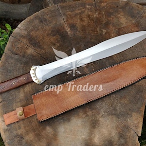 Puede incluir: Un cuchillo grande de acero pulido con mango de madera y detalles de latón, apoyado sobre una superficie de madera. El cuchillo va acompañado de una funda de cuero marrón. El texto "emp Traders" es visible.