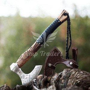 May include: A handcrafted axe with a wooden handle, black leather grip, and a silver-colored head. The axe head features etched designs. A brown leather sheath is included. The axe is displayed outdoors.