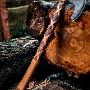 May include: A handcrafted axe with a dark wooden handle wrapped in tan leather. The axe head is made of steel and has a curved blade. The axe rests on a dark wooden log, showcasing the craftsmanship and rustic aesthetic.