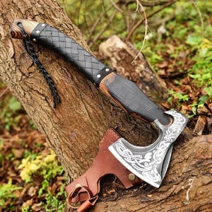 May include: A Viking-style axe with a wooden handle and a black leather-wrapped grip. The axe head features intricate engraved designs and rests on a tree trunk. A brown leather sheath is included.