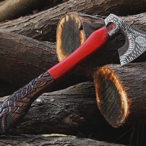 May include: A handcrafted axe with a red handle and a detailed, engraved head. The axe head features intricate patterns and a stylized animal design. The handle is carved with a braided pattern and an eagle head at the base. The axe rests on a pile of wooden logs.