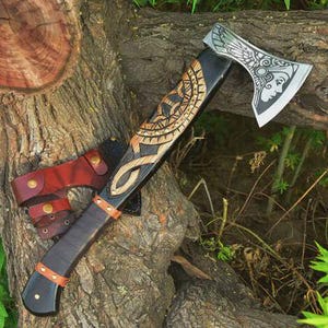 May include: A handcrafted axe with a black handle and a silver blade featuring an engraved woman's face. The axe head has a decorative design. A leather sheath and belt loop are attached. The axe is resting on a tree trunk.