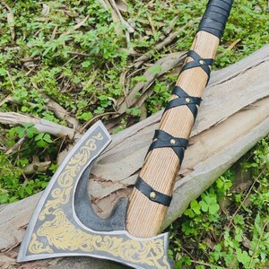 May include: A handcrafted axe with a wooden handle wrapped in black leather. The axe head is gray with gold decorative patterns. The axe is resting on a log with green foliage in the background.