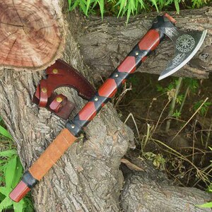 May include: A handcrafted Viking axe with a wooden handle wrapped in brown leather and a black and red diamond pattern. The axe head features an engraved design and a sharp blade. A smaller, matching axe and leather sheath are also present.
