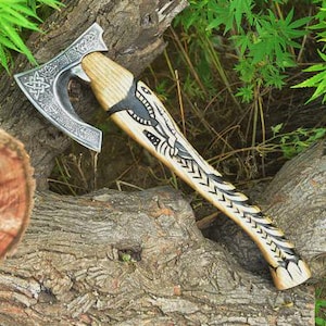 May include: A Viking axe with a silver-colored, engraved blade and a wooden handle. The handle features intricate black carvings and a light brown finish. The axe is resting on a weathered, textured log, showcasing its craftsmanship and design.