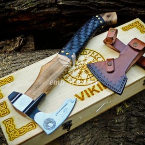 May include: A handcrafted Viking axe with a wooden handle, black leather grip, and a silver-colored blade. The axe rests on a wooden box with Viking symbols and the word "VIKING" engraved on it. A brown leather sheath is also present.