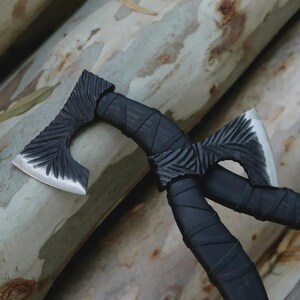 May include: Two small, decorative axes with black handles and intricately carved heads. The axe heads have a silver finish. The axes are crossed, with the handles wrapped in a textured material.