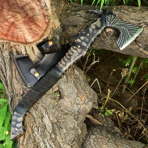 May include: A handcrafted axe with a patterned wooden handle and a black leather sheath. The axe head is silver with a decorative design. The handle is wrapped in black leather. The axe is resting on a tree trunk.