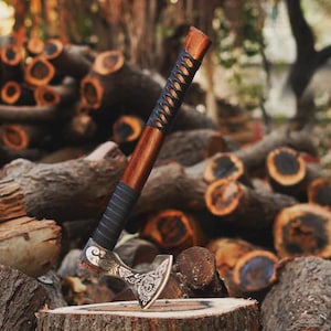 May include: A handcrafted axe with a wooden handle and a decorative metal head. The handle is wrapped with black leather and features a dark brown wood grain. The axe is resting on a tree stump in front of a pile of logs.