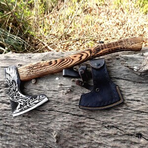 May include: Axe with a wooden handle and a black and silver blade, resting on a log. The handle has carved details, and a black leather sheath is included. The axe blade features a decorative design.