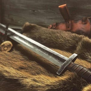 May include: A medieval-style sword with a silver blade and a leather-wrapped handle rests on a bed of brown fur. A brown leather sheath and a wooden handle are also visible, suggesting a historical or fantasy theme.