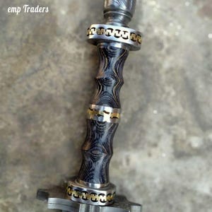 Op de afbeelding: Close-up van een decoratieve dolk met een patroon op het lemmet en een houten handvat met textuur. De handgreep is versierd met zilver- en goudkleurige metalen ringen. De tekst "emp Traders" is zichtbaar linksboven.